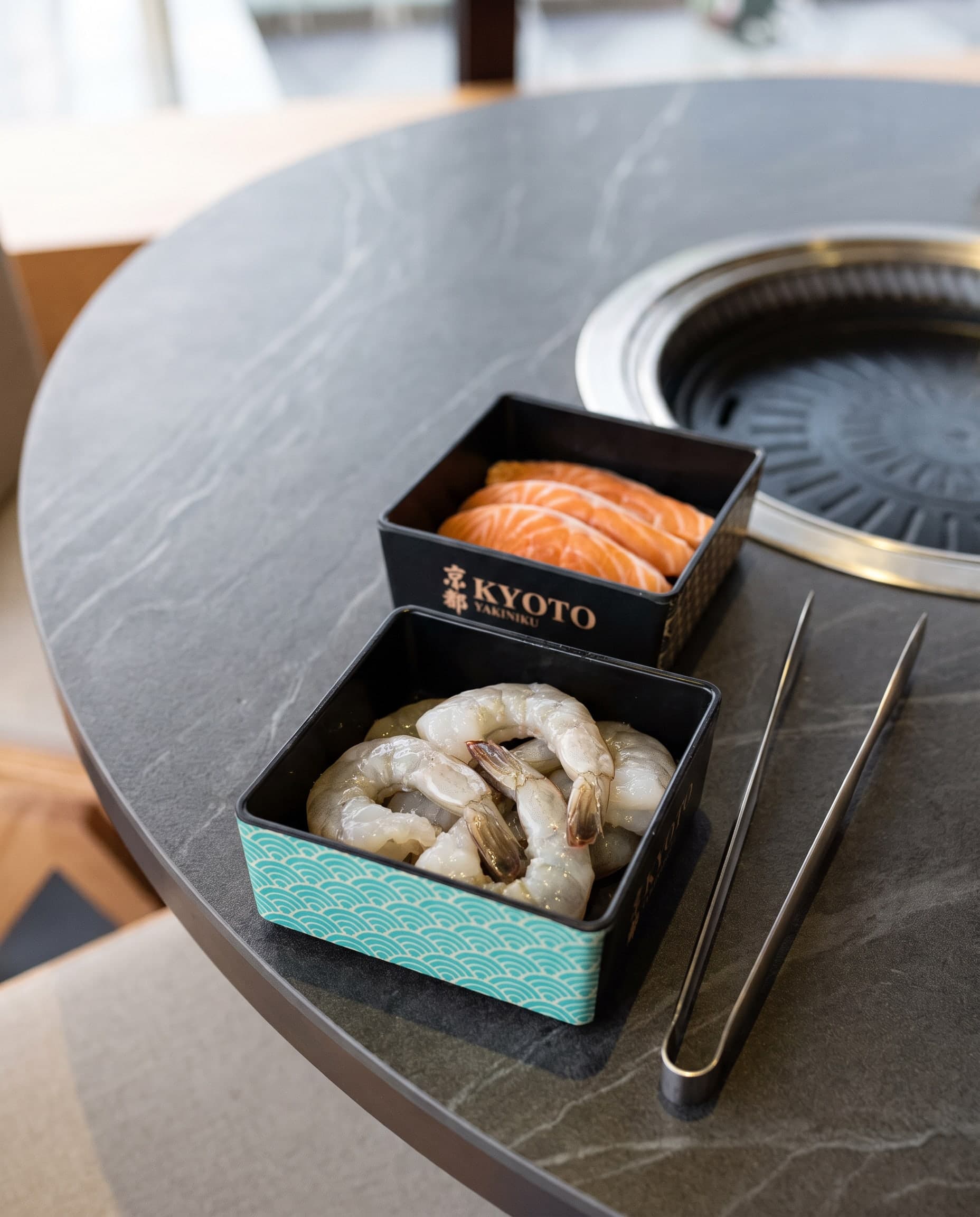 Boxen mit rohen Garnelen und Lachsscheiben neben dem Tischgrill, daneben eine Grillzange