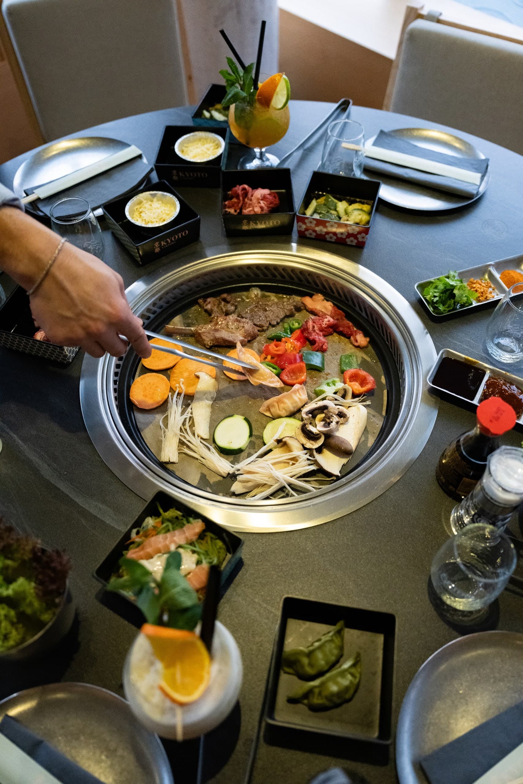 Tischgrill von oben mit Fleisch, Gemüse und Pilzen, rundherum Beilagen und Getränke